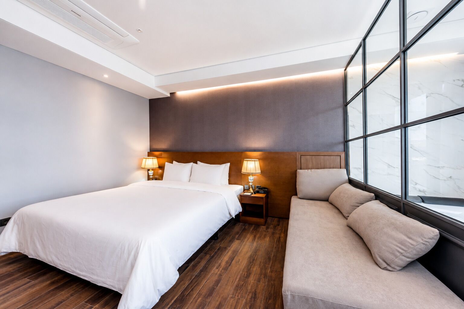A hotel room with a large bed, a sofa, a nightstand with lamps, and a large window with a view.