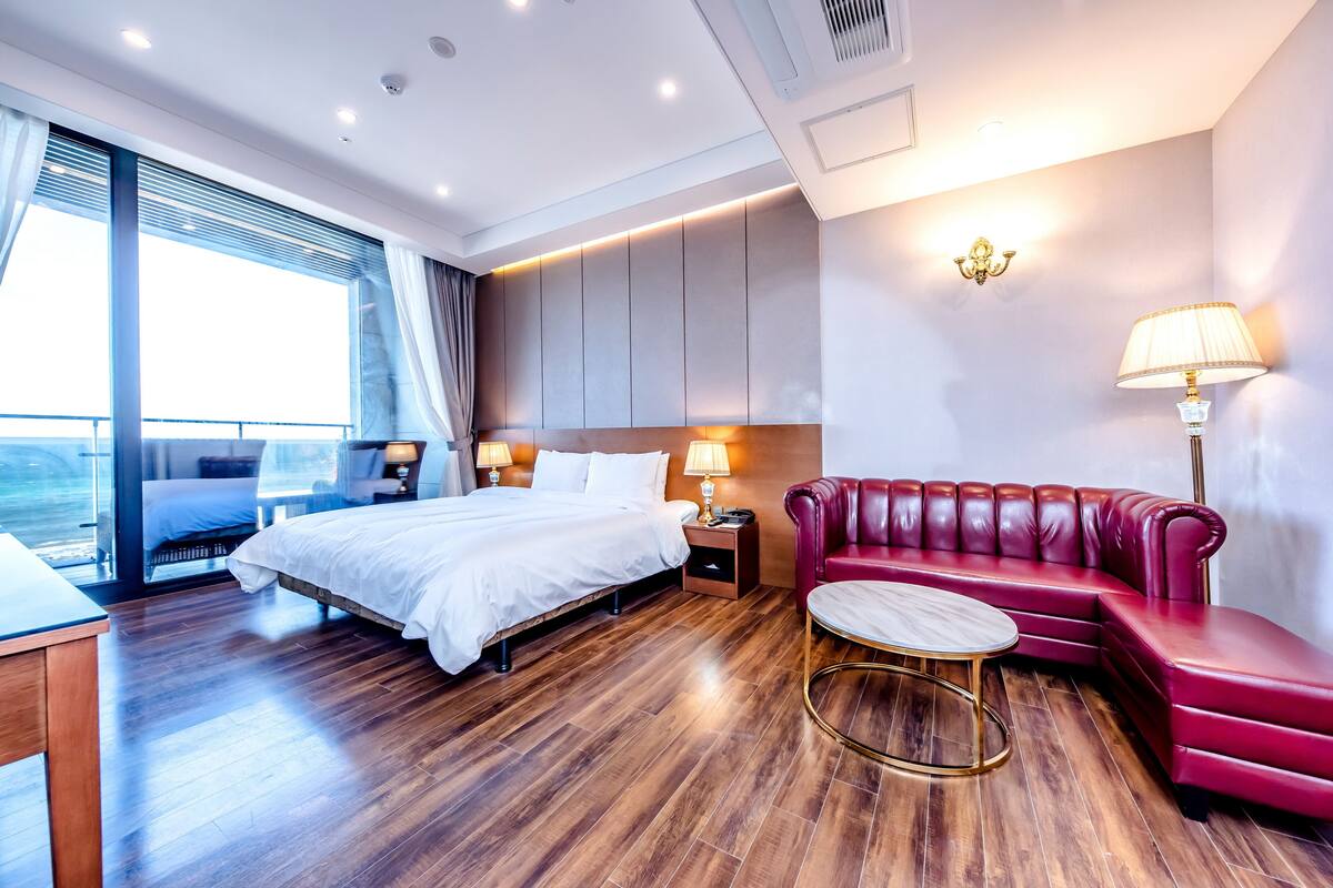 Une chambre d’hôtel moderne avec un grand lit, un canapé en cuir rouge, une table basse ronde et une vue sur la mer.