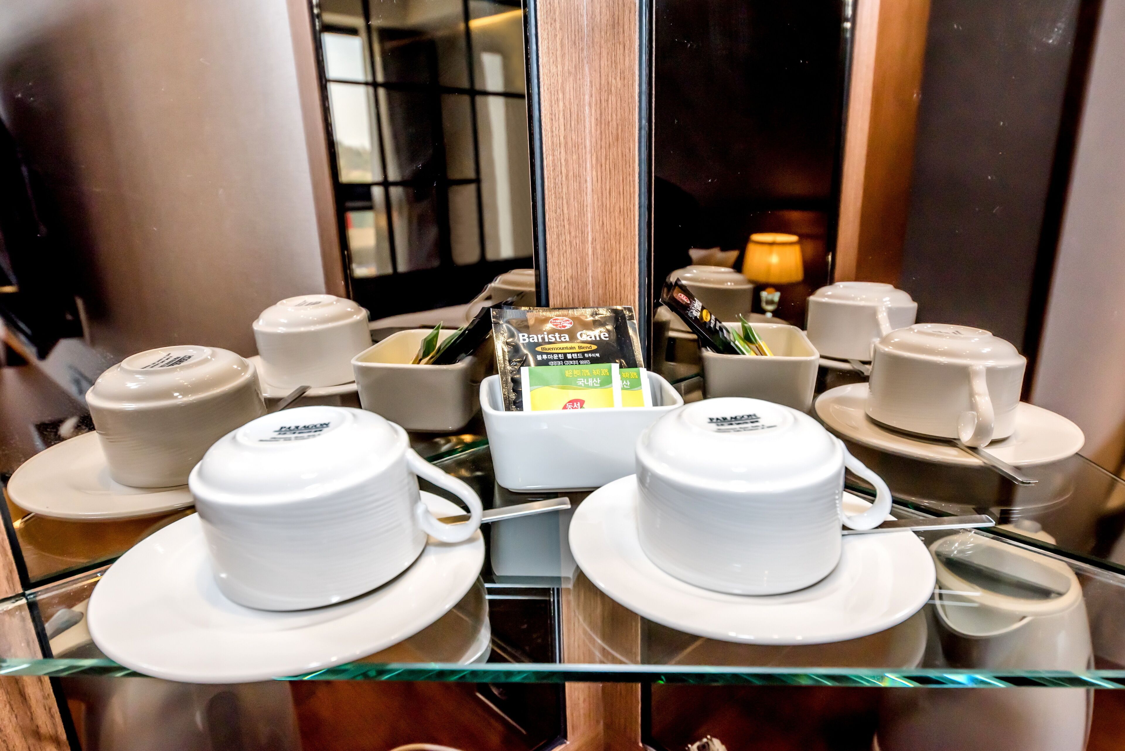 Habitación de hotel con una mesa de vidrio que muestra tazas y platillos blancos, una máquina de café tipo barista y un paquete de café.