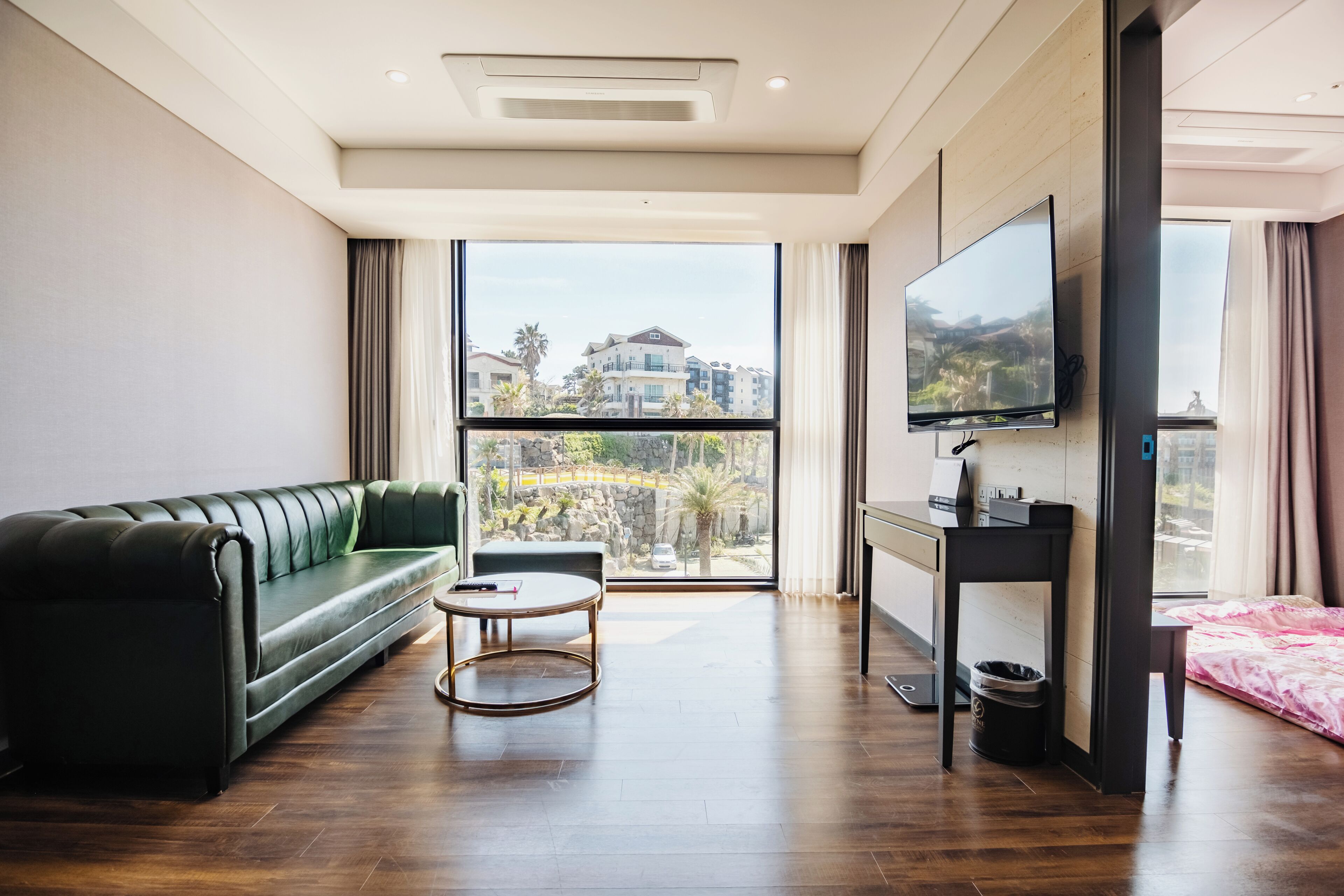 Un soggiorno moderno con un divano verde, un tavolino rotondo, una TV a parete e un'ampia finestra con vista su edifici e palme.