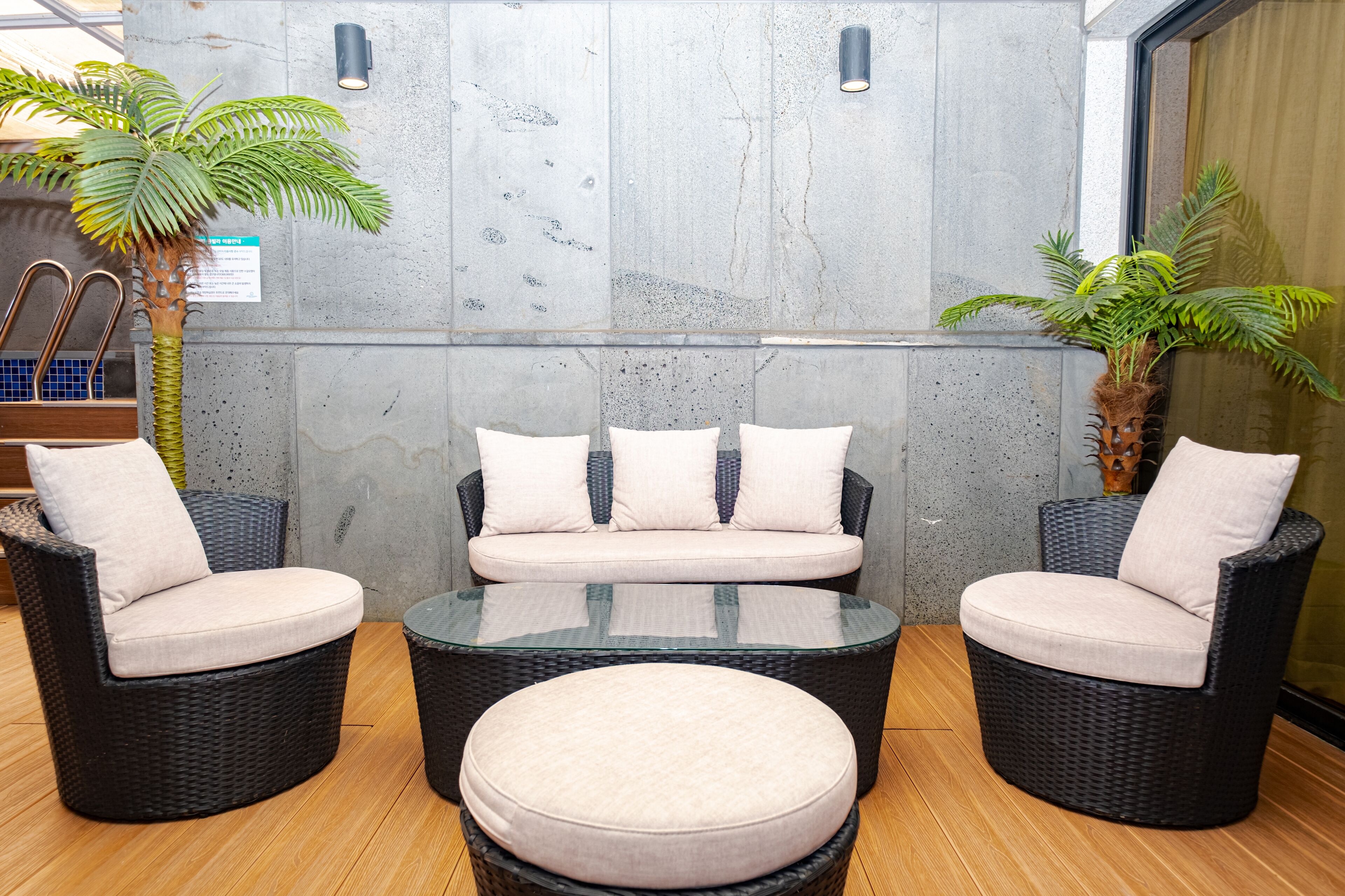 A seating area with wicker furniture and a glass-top table, set against a concrete wall with a plant on the left and a window on the right.