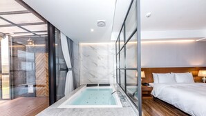 A modern hotel room with a marble bathtub, a wooden floor, a large bed with white linens, and a glass shower enclosure.