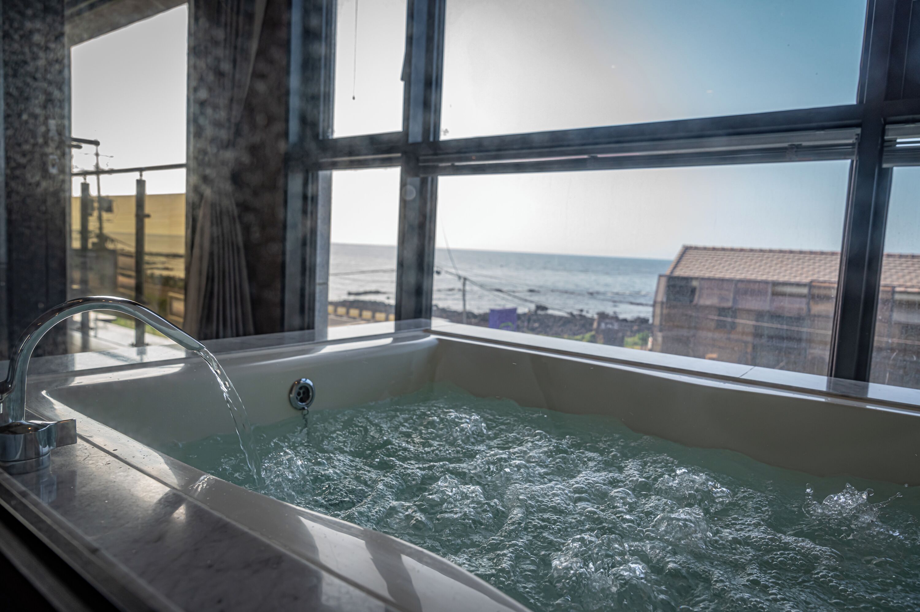 A bathtub with running water, large windows, and a view of the ocean.