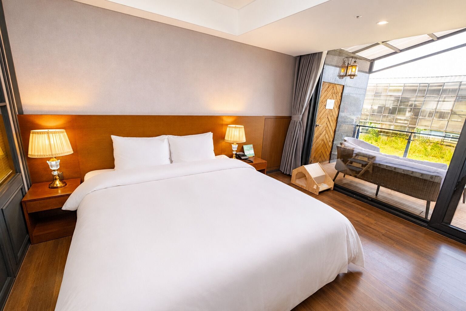 A hotel room with a large bed, two bedside lamps, a small table, a chair, and a balcony with a view.