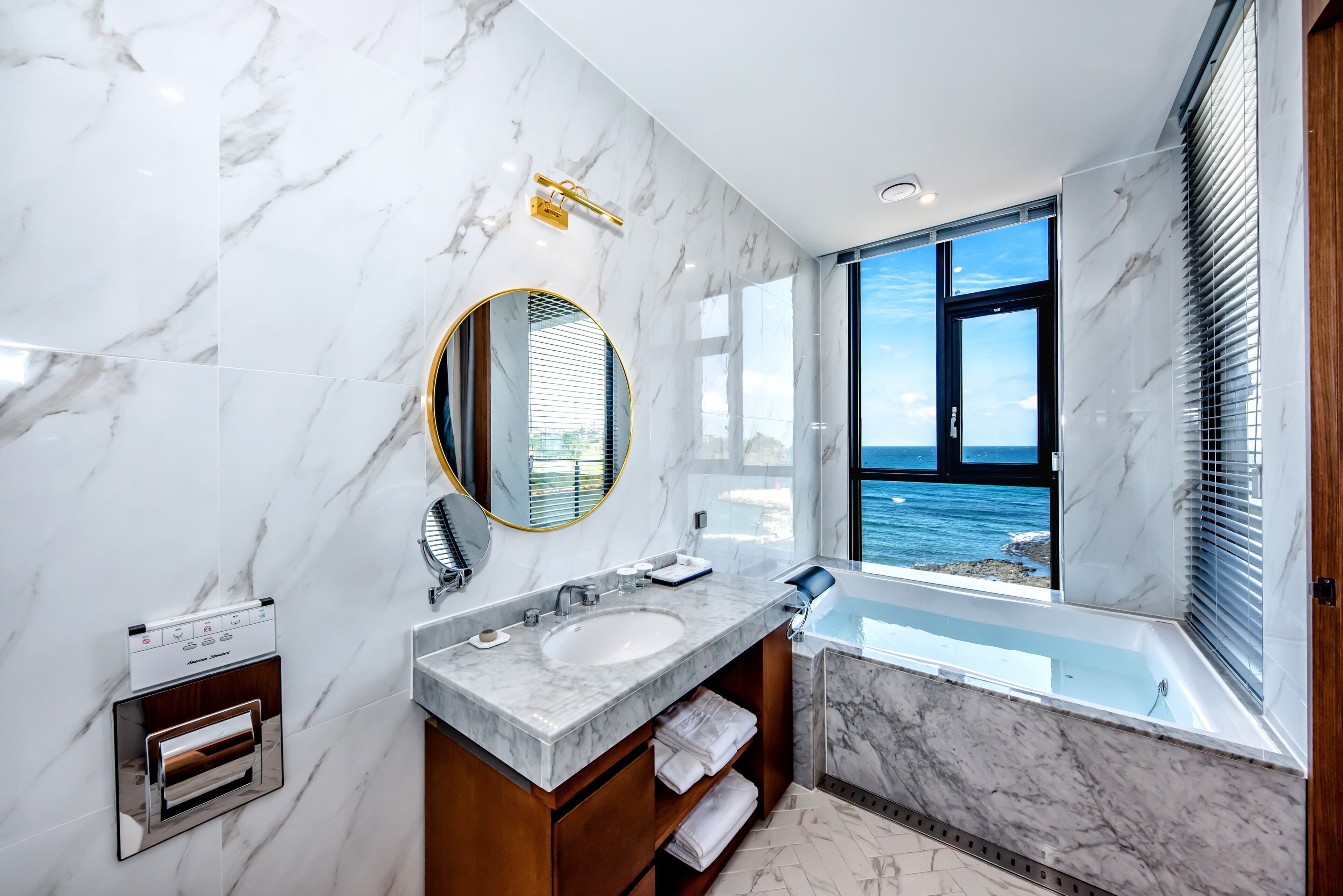 A modern bathroom with a marble vanity, a built-in oven, a large mirror, and a bathtub with a view of the sea.