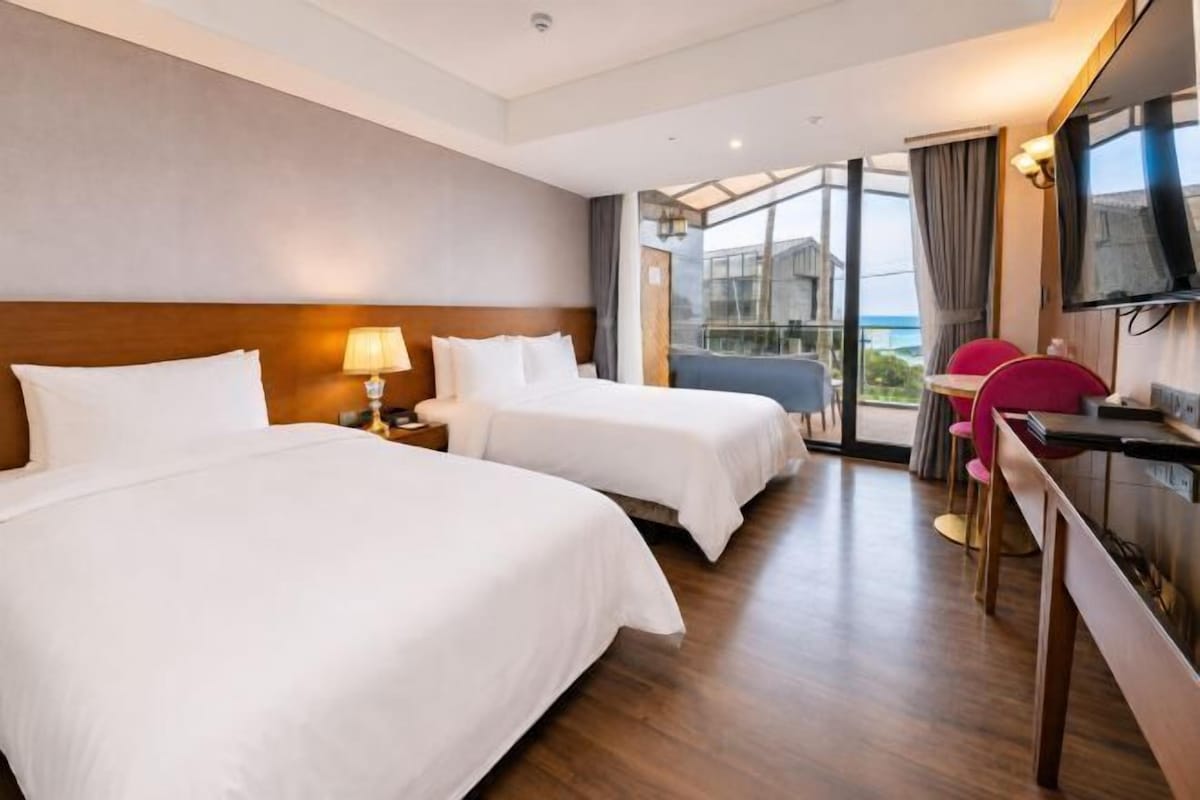 A hotel room with two beds, a desk, a chair, a TV, and a view of the outdoors.
