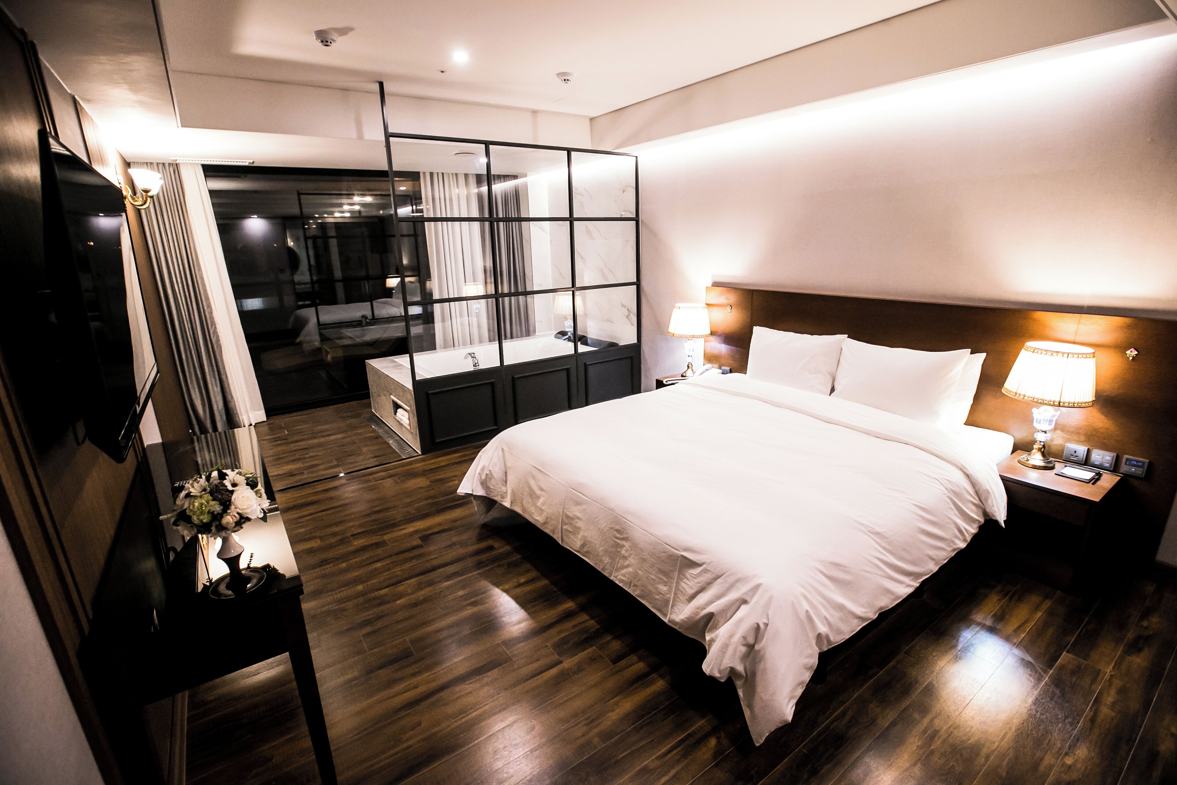Una habitación de hotel moderna con una cama grande, un tocador con espejo, una mesita de noche con lámpara y vistas a un balcón.