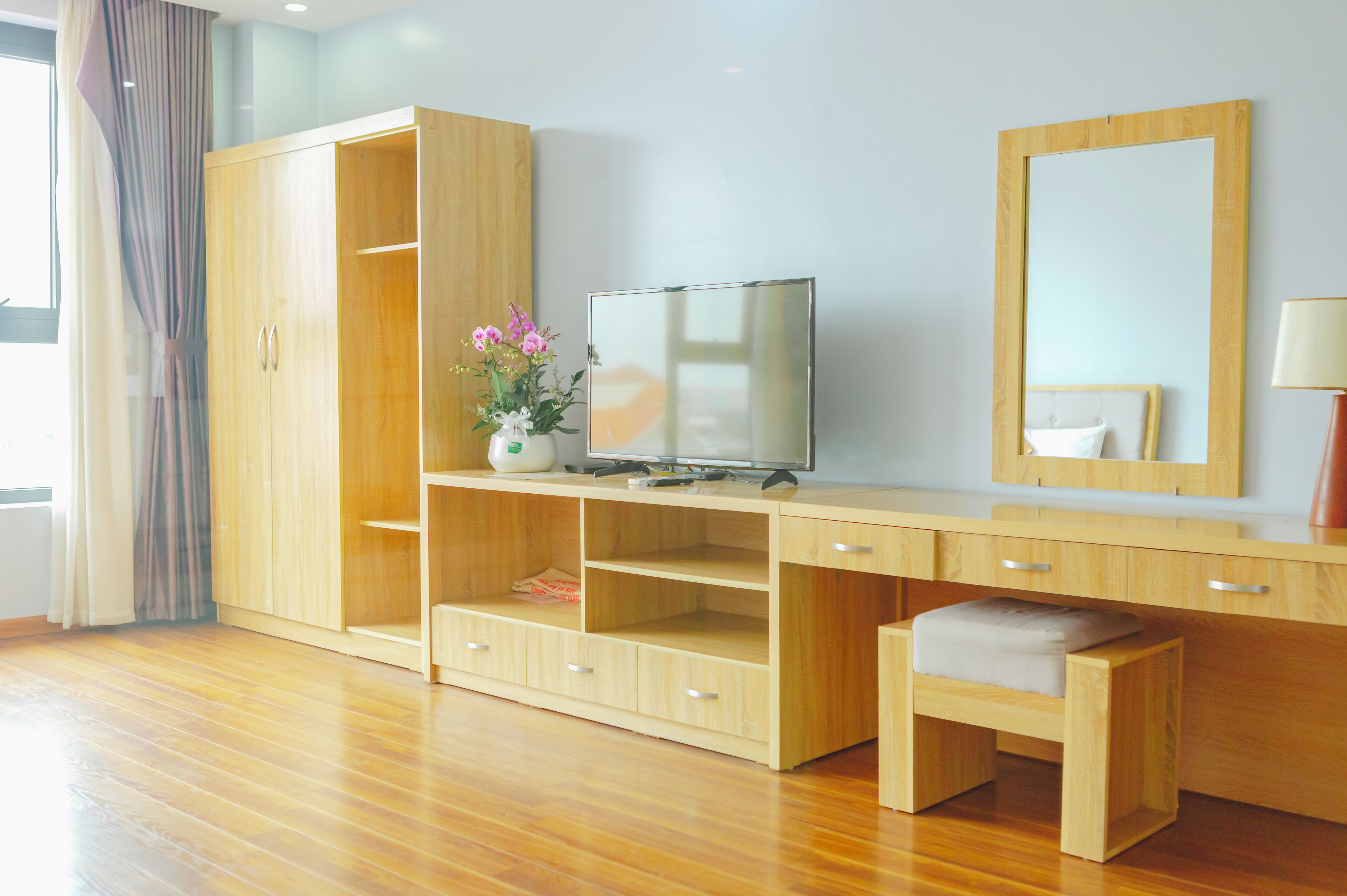 A hotel room with a wooden wardrobe, a TV on a wooden stand, a mirror, and a small bench.