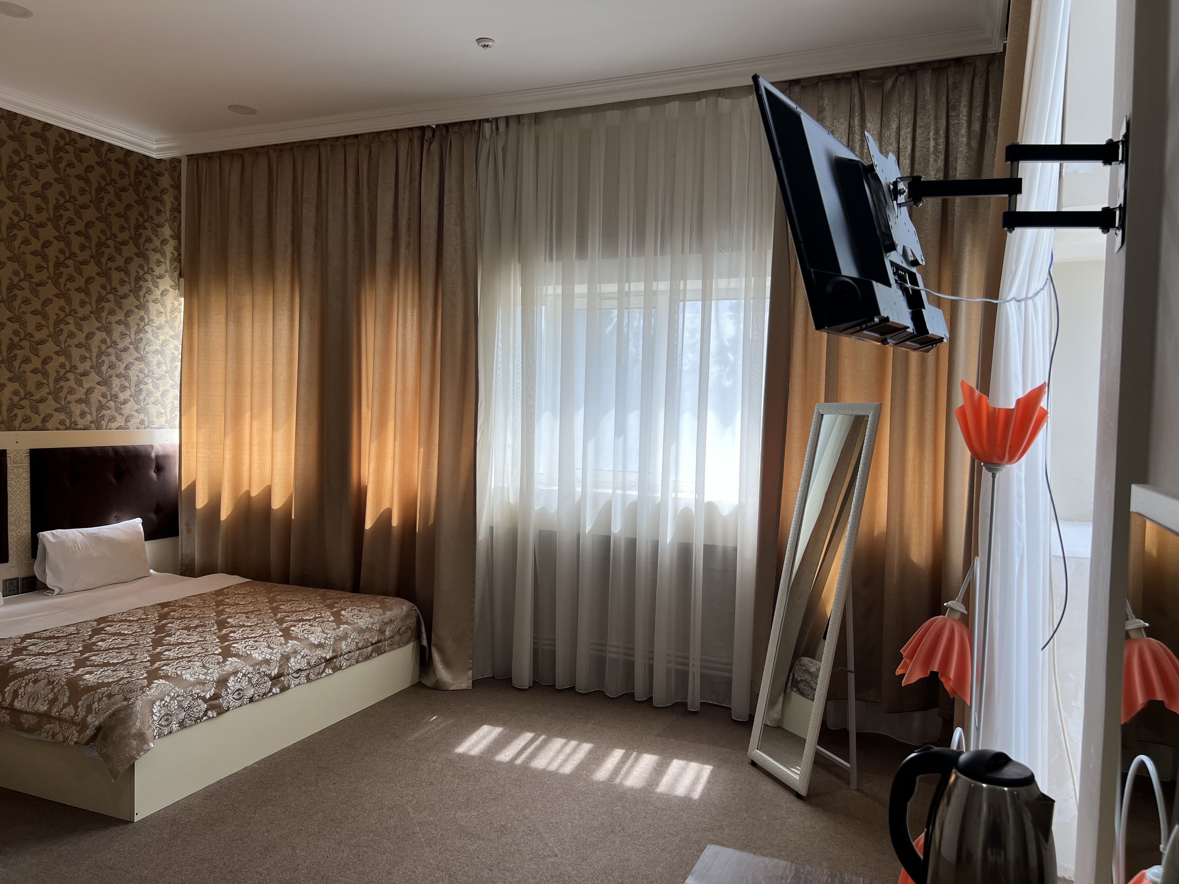 A hotel room with a bed, a flat-screen TV mounted on the wall, a large mirror, and a kettle on the floor.