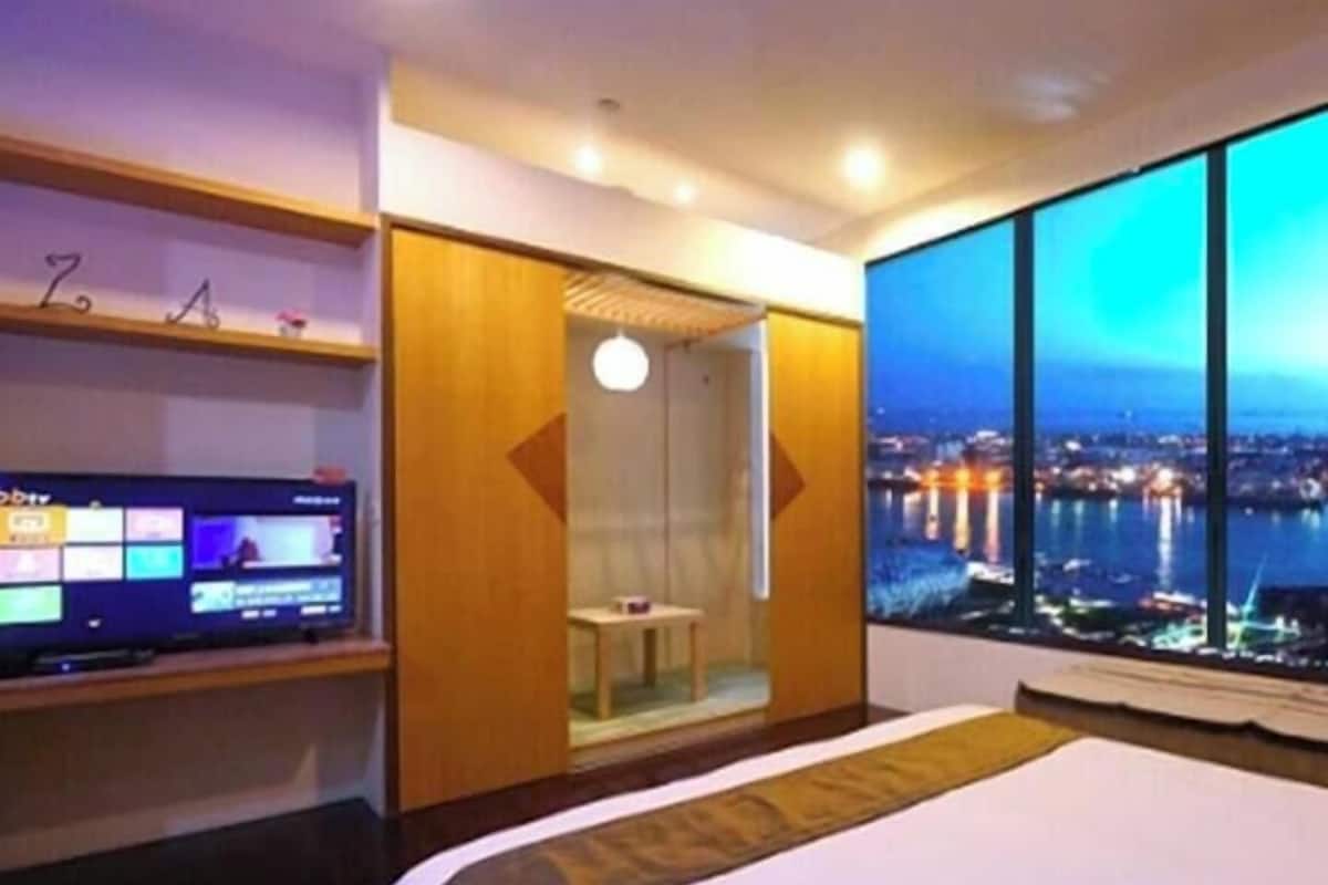 Habitación de hotel con cama, televisor, una mesita, zona de ducha y un ventanal con vistas a la ciudad.