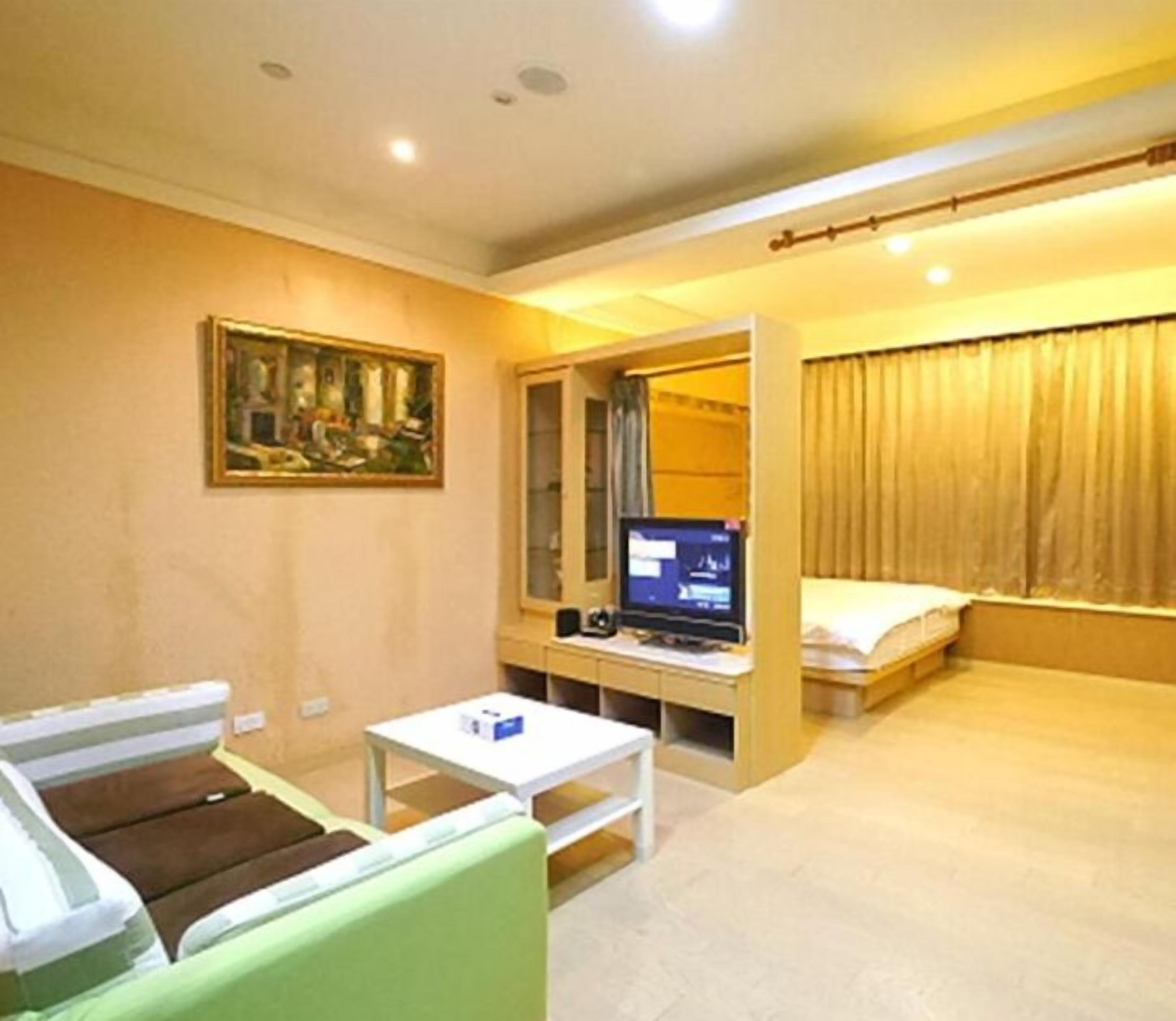 A small, well-lit room with a bed, a desk with a computer, a chair, and a painting on the wall.