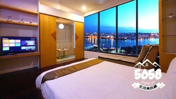 Habitación de hotel con una cama grande, un televisor sobre un mueble, una puerta de cristal que da a un balcón y vistas a la ciudad al anochecer.