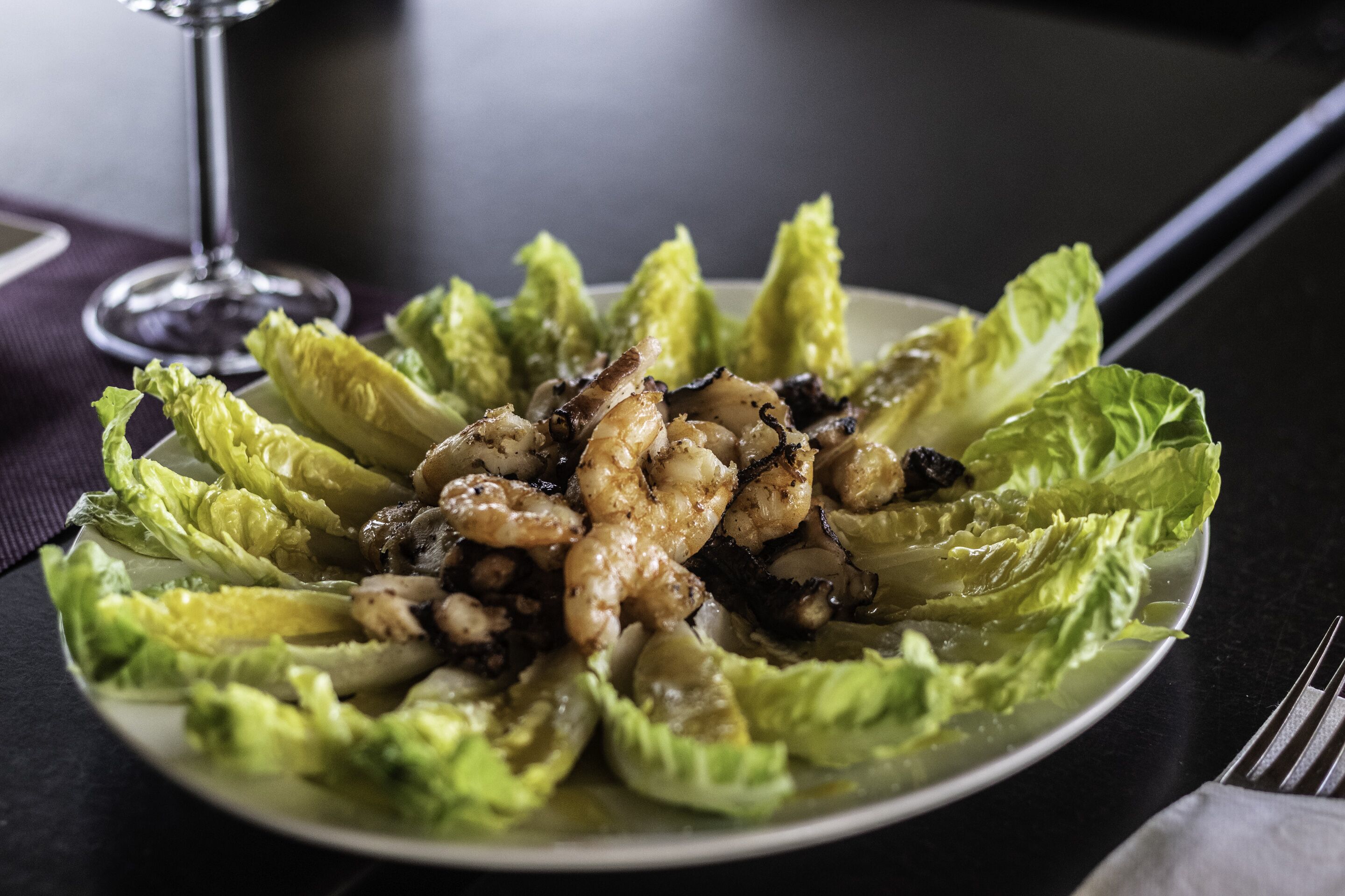 A plate of food with grilled shrimp and mushrooms on lettuce leaves, accompanied by a glass of wine.