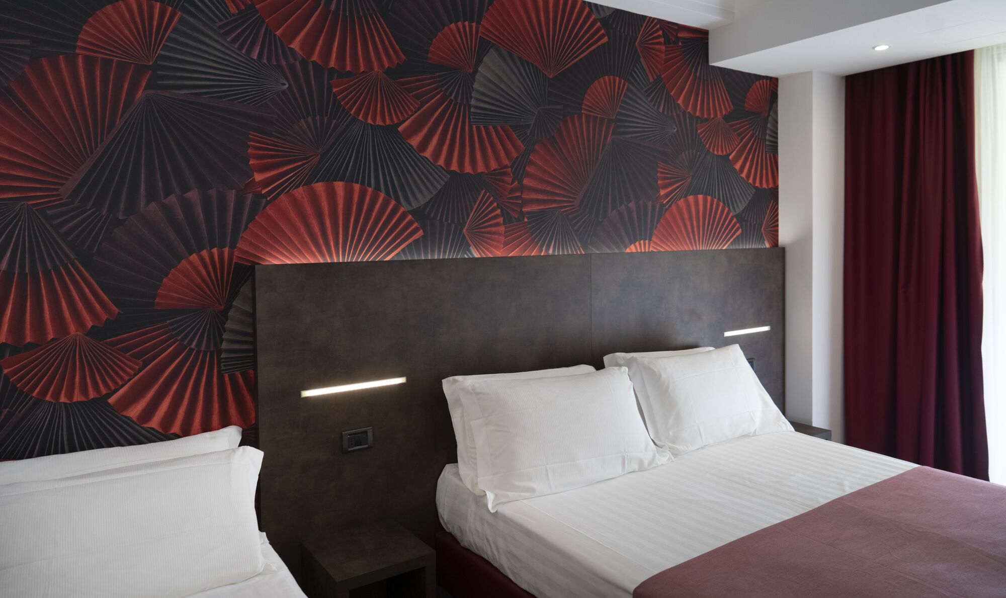 Una habitación de hotel con una cama grande, dos mesitas de noche y una pared con un estampado de abanicos rojo y negro.