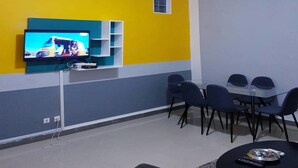 Sala de estar moderna com televisão de tela plana fixada em uma parede verde-água, uma janela pequena e uma área de jantar com cadeiras e mesa.