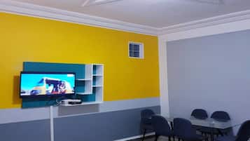 Un salon moderne avec un téléviseur à écran plat fixé sur un mur de couleur turquoise, une petite fenêtre et un coin repas comprenant des chaises et une table.