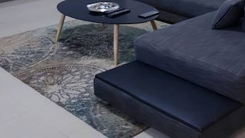 Un canapé gris foncé accompagné d’un pouf assorti, une petite table d’appoint ronde sur laquelle reposent une télécommande et un objet décoratif, et un tapis à motifs sur le sol.