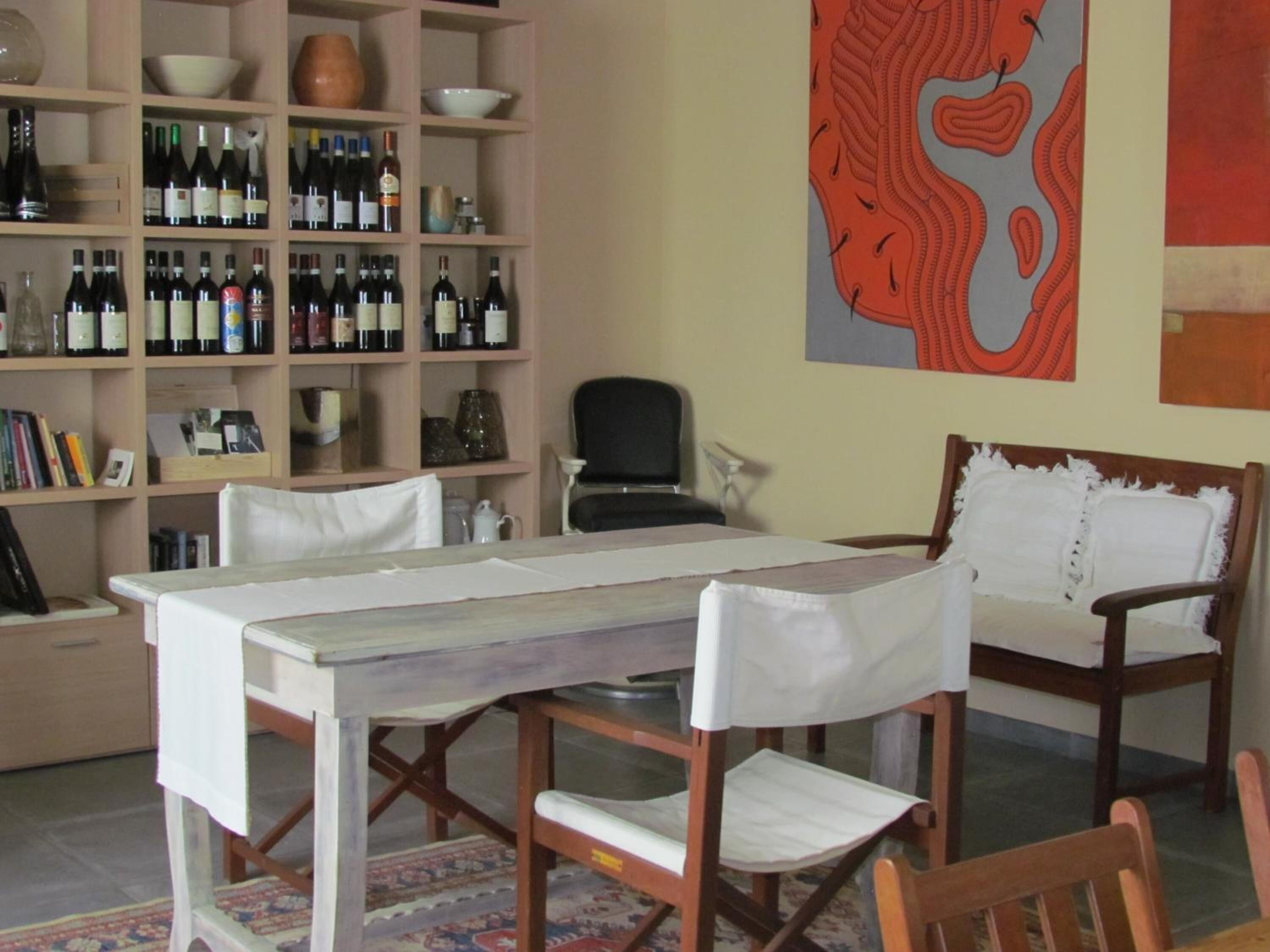 Een eetgedeelte met een tafel en stoelen, een boekenkast met wijnflessen, en een groot abstract schilderij aan de muur.