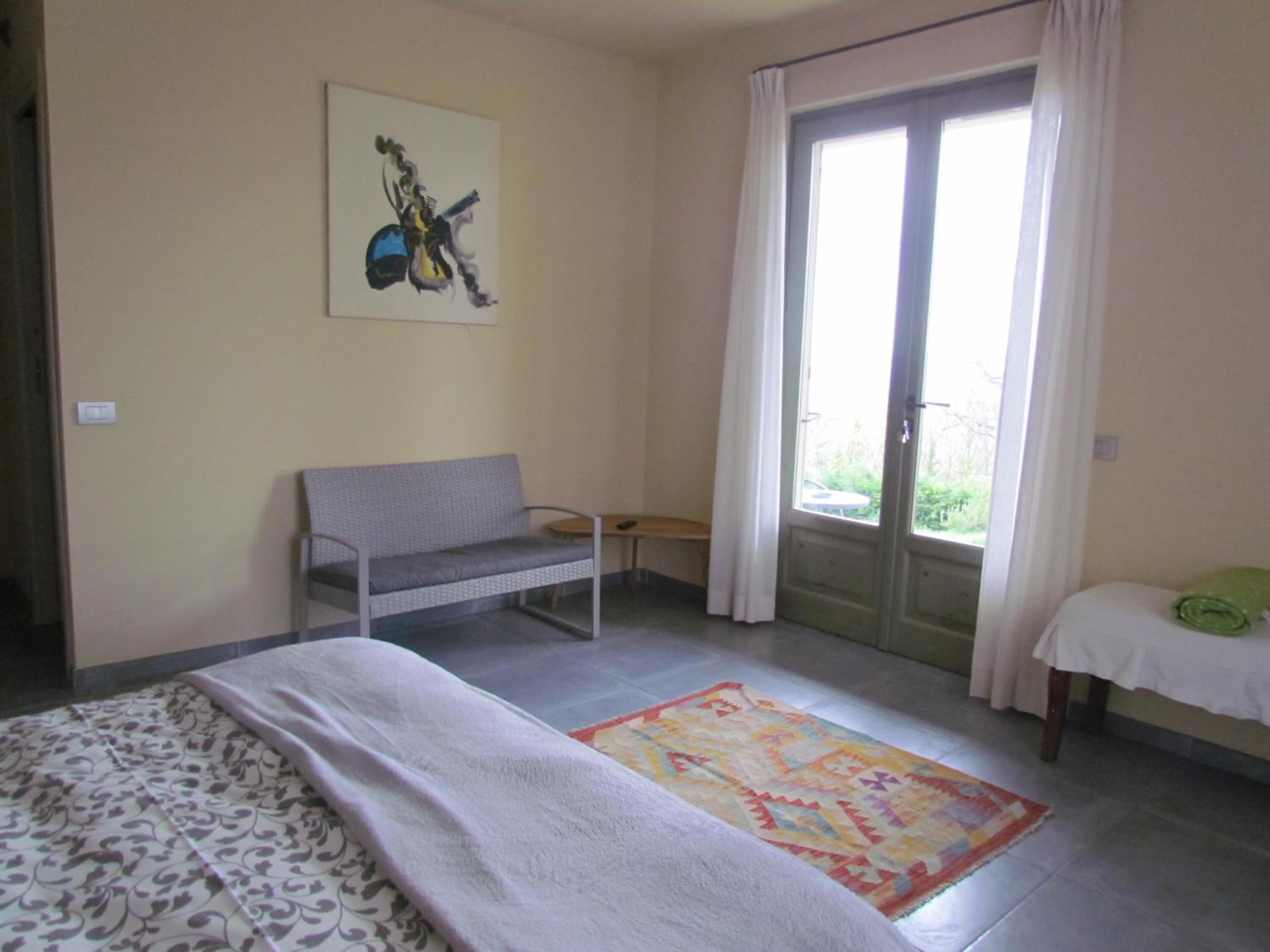 Een slaapkamer met een bed, een bankje, een kleurrijk vloerkleed en een schilderij aan de muur.