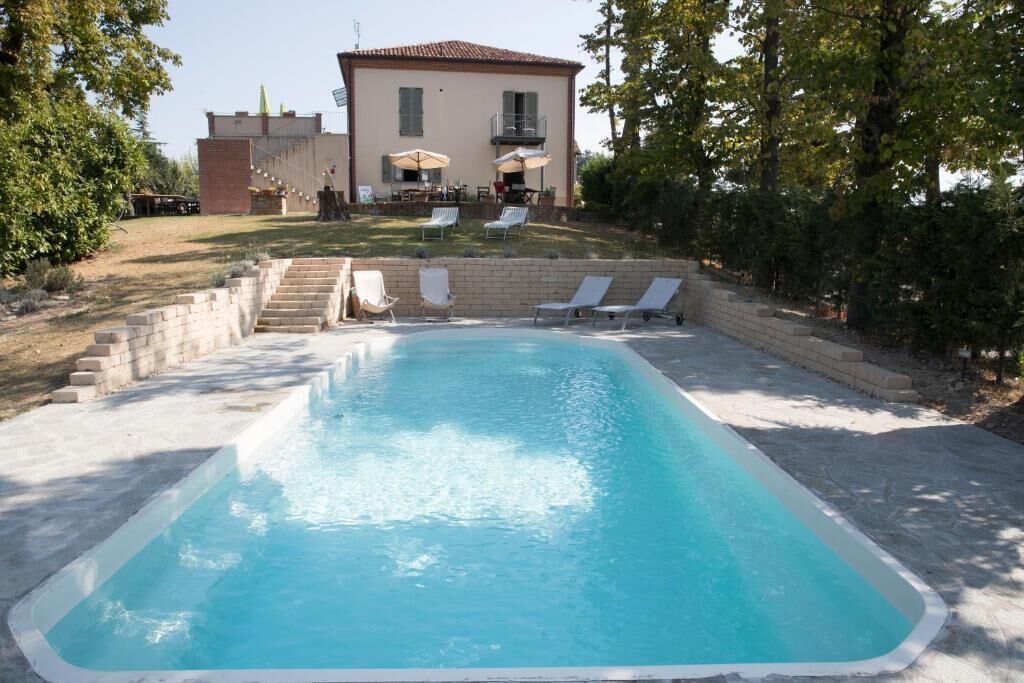 Una piscina rettangolare con superficie d'acqua cristallina, circondata da una platea in cemento. Sulla platea si trovano poltroncine da relax bianche e ombrelloni, e sullo sfondo un edificio con tetto rosso e finestre.