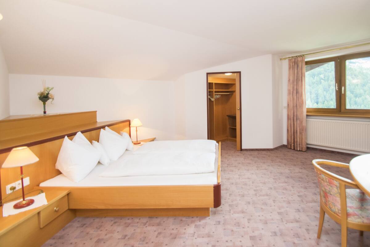Quarto de hotel com cama de madeira, luminárias de cabeceira, uma mesa pequena e uma janela com cortinas.