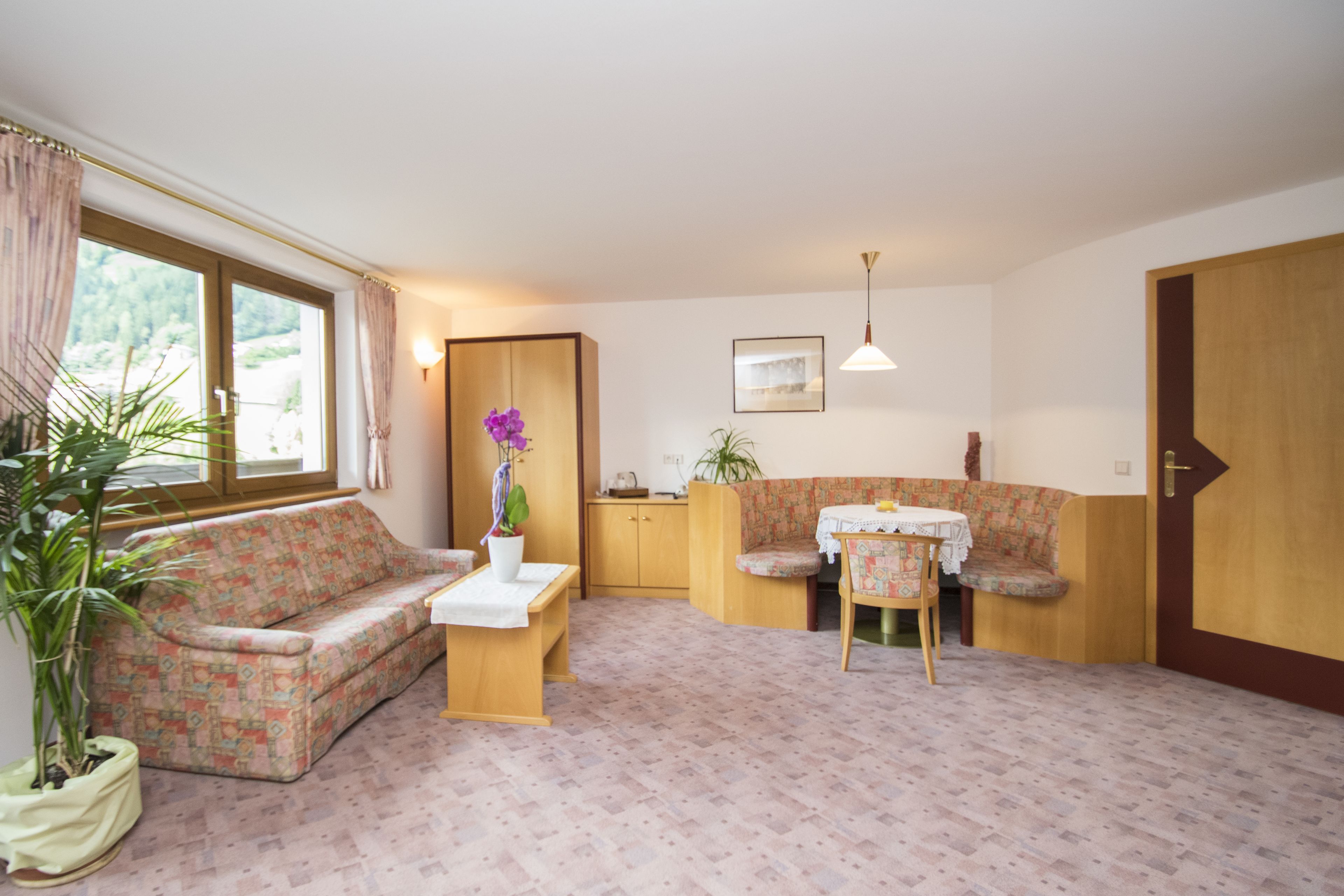 Una habitación acogedora con un sofá floral, una pequeña mesa de comedor con sillas, una planta en maceta y una puerta de madera.