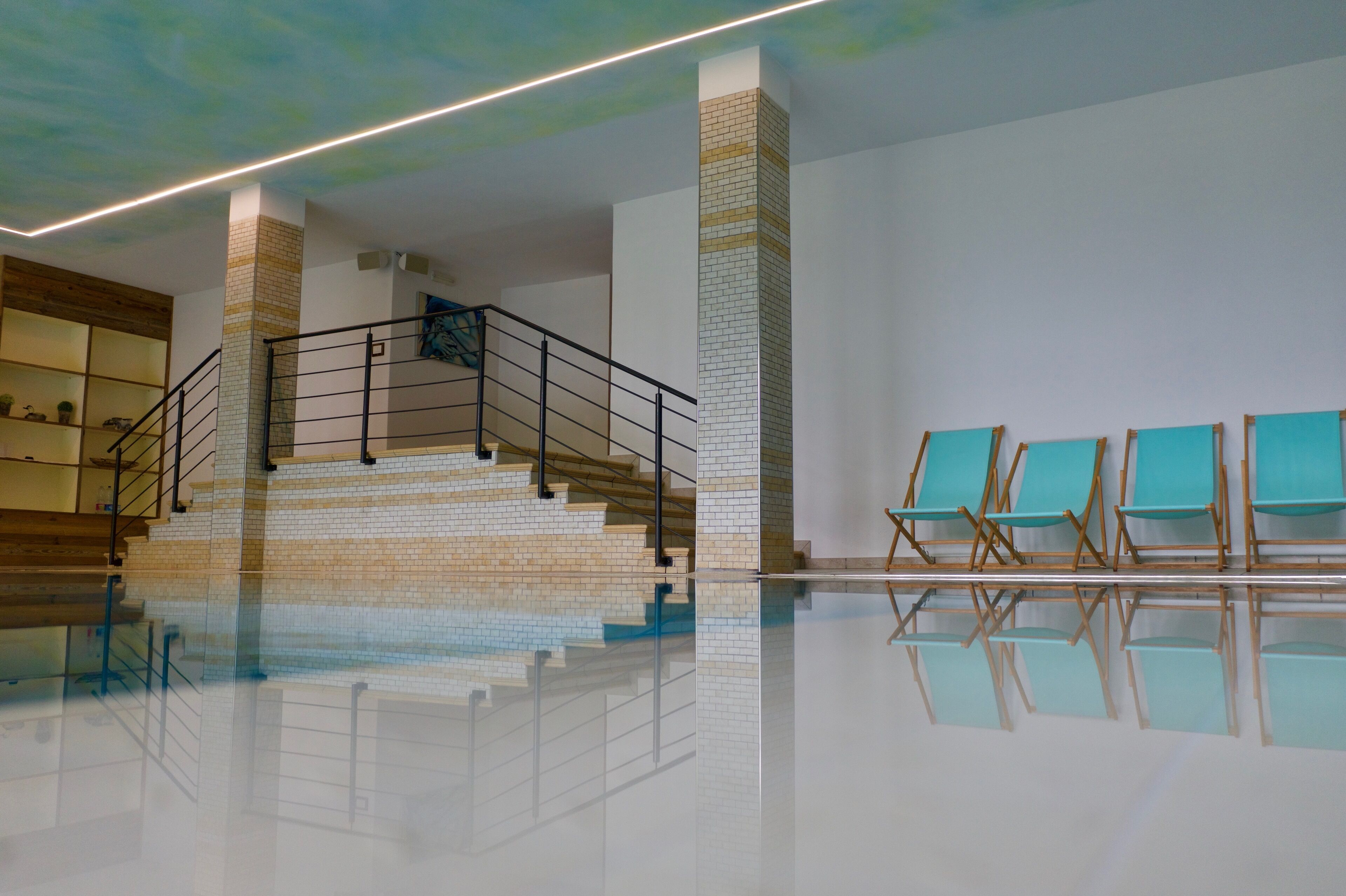 Un área de piscina interior espaciosa con una escalera, un muro de azulejos y varias sillas de descanso.