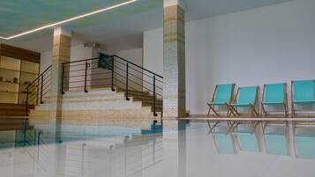 Un área de piscina interior espaciosa con una escalera, un muro de azulejos y varias sillas de descanso.