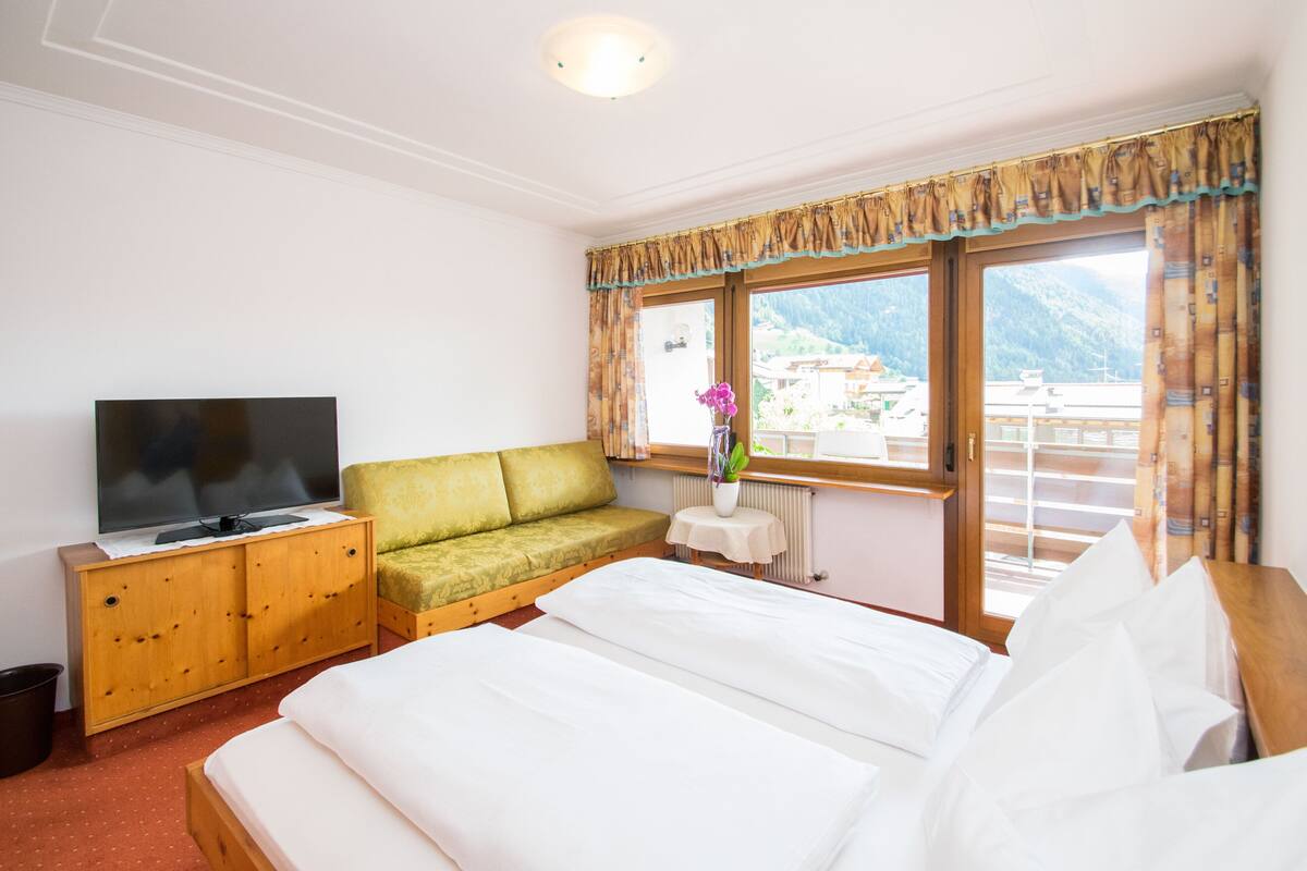Quarto de hotel com uma cama, um sofá verde, uma televisão, uma mesa pequena com um vaso e uma janela com cortinas.