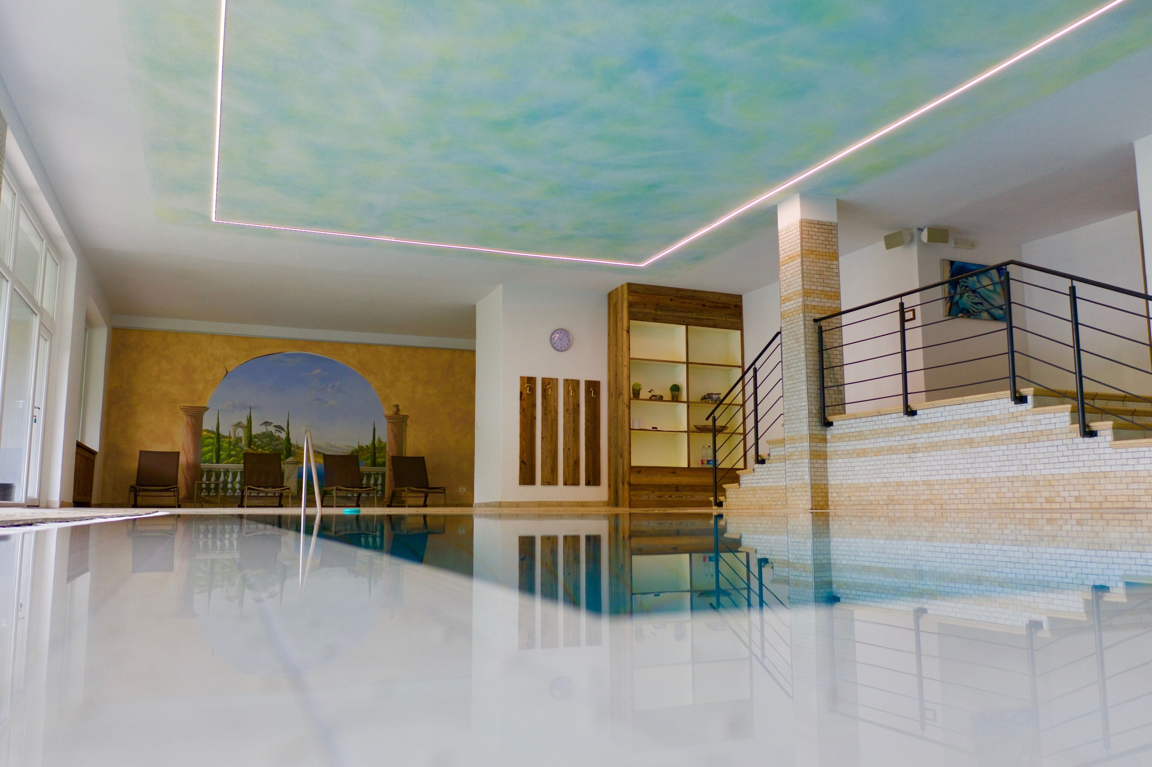 A modern indoor swimming pool with a clear ceiling, a wooden staircase, and a mural on the wall.