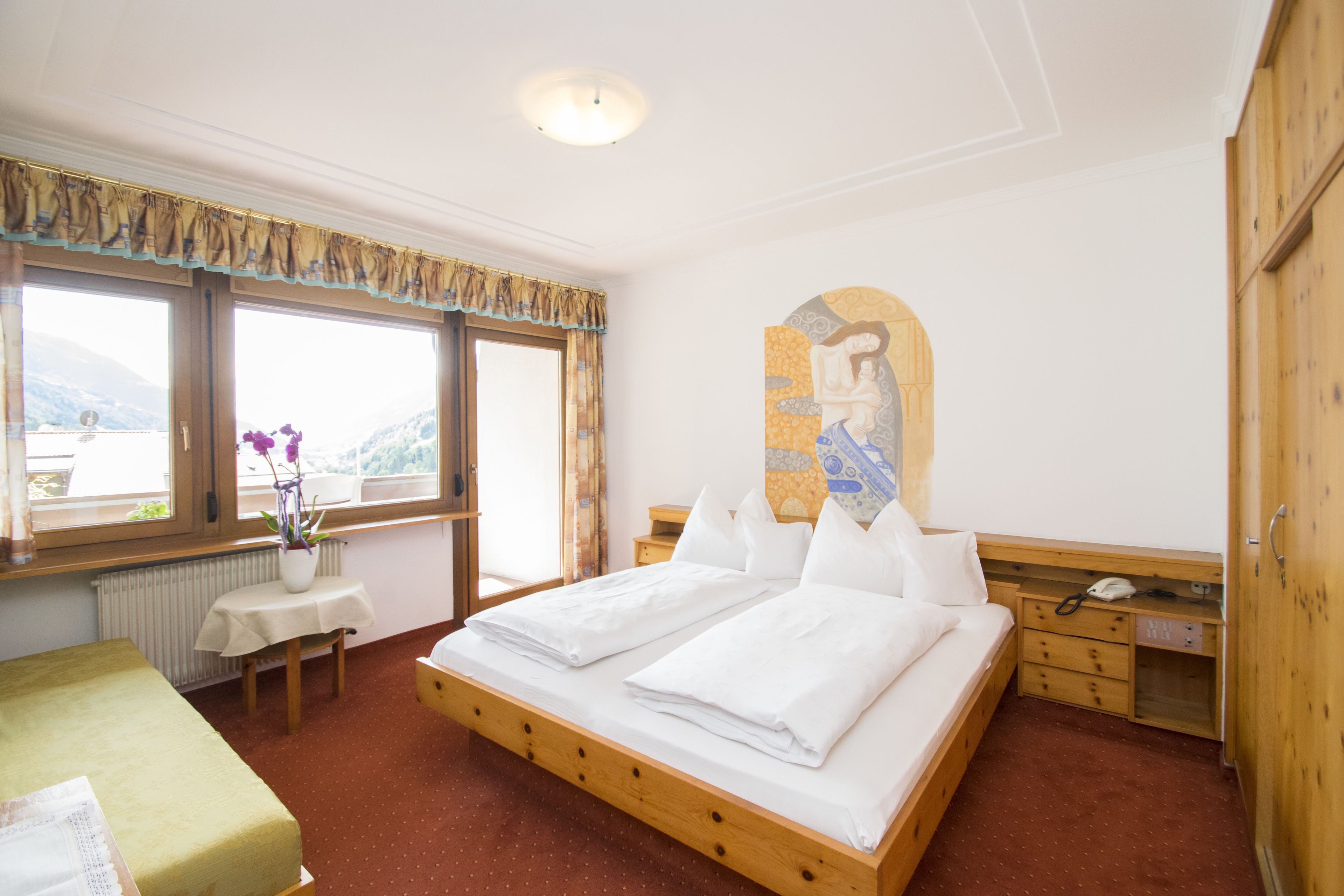 Un dormitorio con cama de madera, una mesita con un jarrón de flores, una ventana con vistas a montañas y un armario de madera.