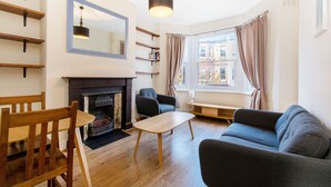 A living room with a fireplace, wooden furniture, a sofa, and a window with curtains. - Simon Apartment - London Launchpad (London)