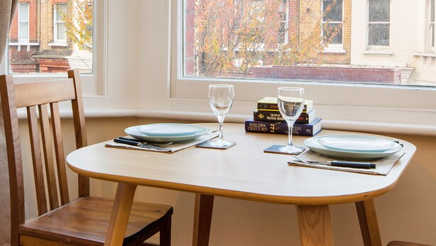 A dining area with a table set for two, a chair, and a view of a cityscape through large windows. - Simon Apartment - London Launchpad (London)