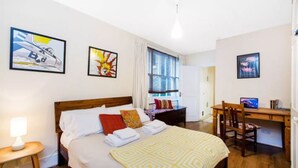 A hotel room with a bed, a desk, and a chair. There are paintings on the walls and a window with curtains. - Simon Apartment - London Launchpad (London)