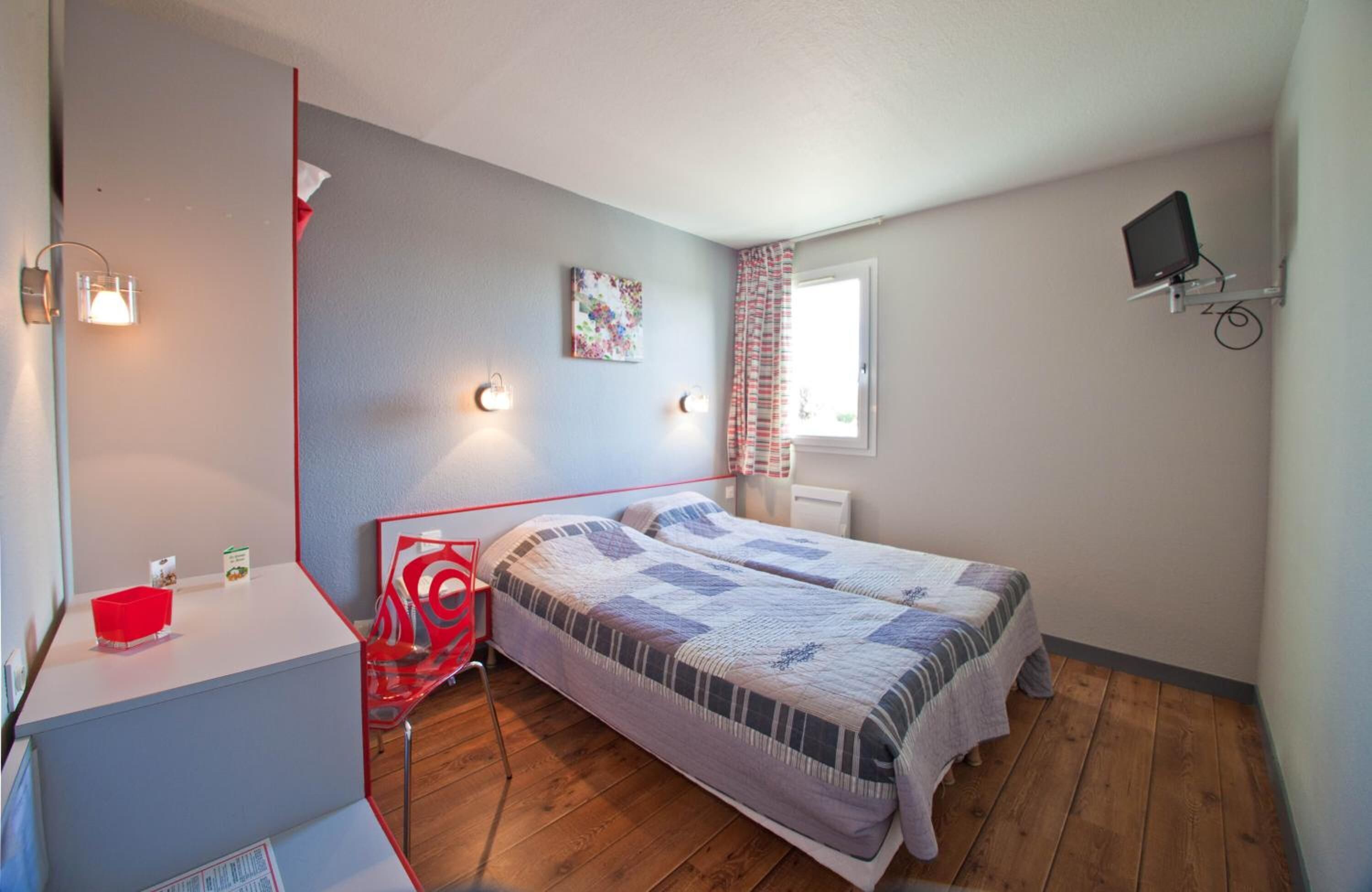 Ein kleines, kompaktes Zimmer mit einem Einzelbett, einem Schreibtisch mit Stuhl, einem kleinen Tisch und einem an der Wand befestigten Fernseher.