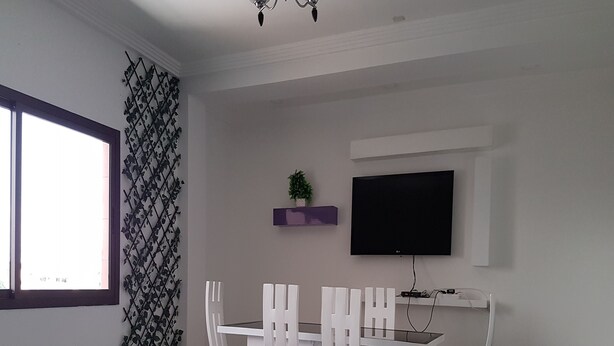 A dining area with a white table and chairs, a flat-screen TV mounted on the wall, and a potted plant on a shelf.