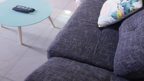 A bedroom with a bed covered in a magazine-style duvet, a grey sofa with a floral pillow, and a small round table with a remote control.