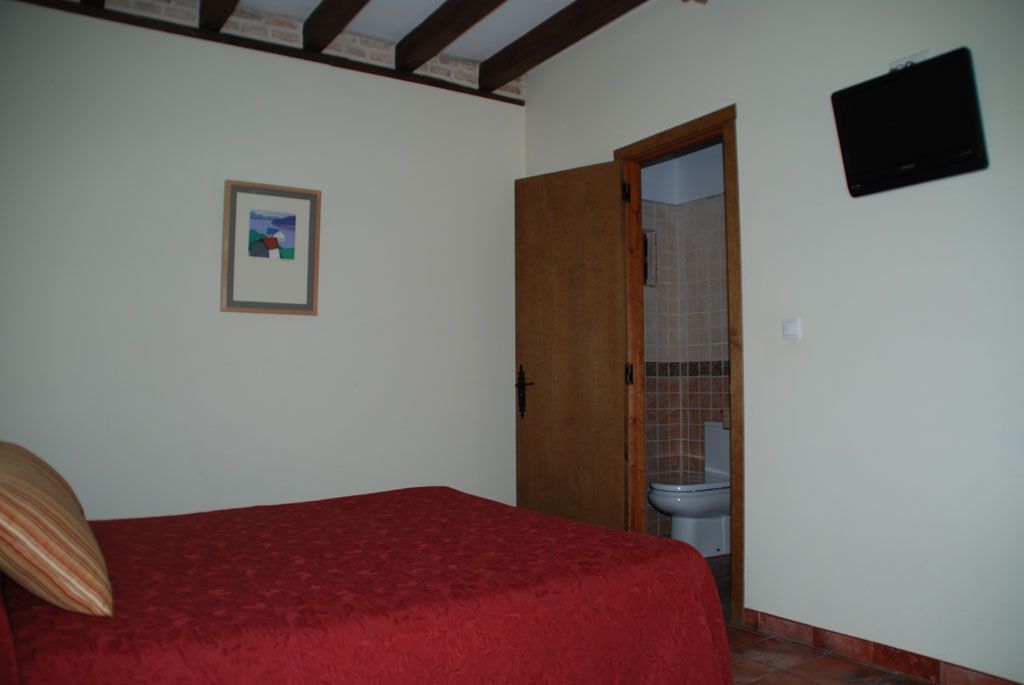 Een slaapkamer met een rode sprei, een houten deur naar een badkamer en een ingelijst beeld aan de muur.