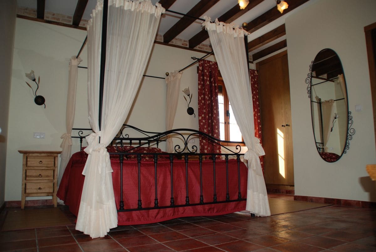 A four-poster bed with red bedding, a wooden nightstand, a mirror, and a red curtain.