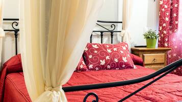 Una cama con colcha y almohadas rojas, una mesita de noche de madera con una planta verde, y un cabecero de metal negro.