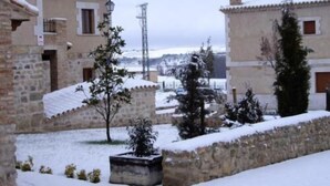 Um pátio coberto de neve com edifícios de pedra, uma pequena árvore e um muro também coberto de neve.