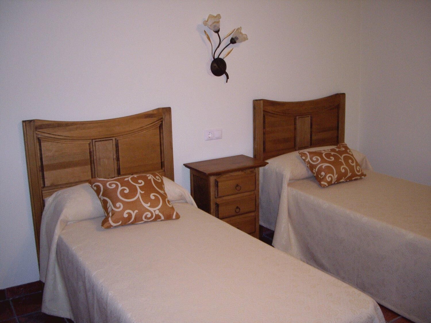 Two single beds with wooden headboards, a small bedside table, and a wall-mounted light fixture with flowers.