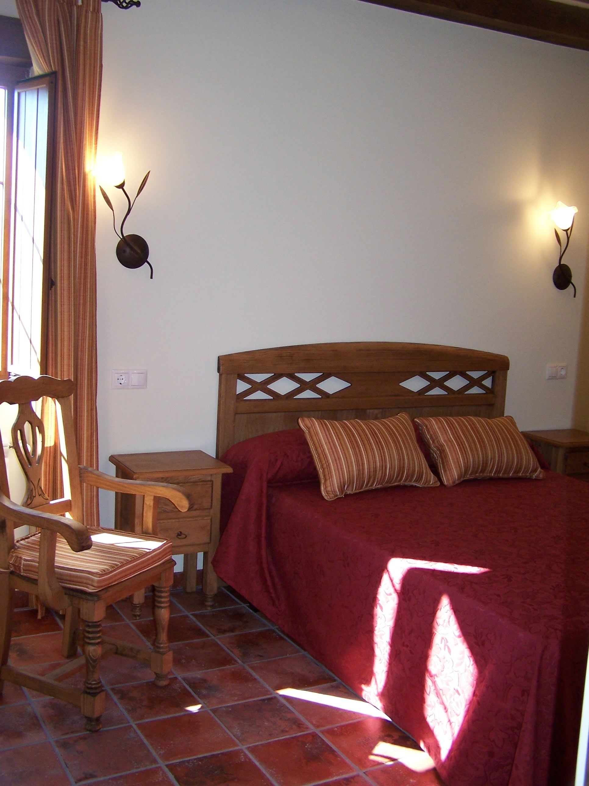Un dormitorio con cama y silla de madera, una mesita de noche y una lámpara fijada a la pared.