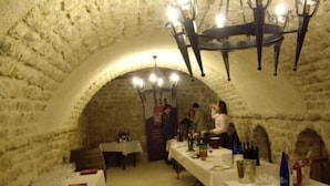 Uma adega subterrânea com paredes de pedra, mesas preparadas para refeições e um candelabro.