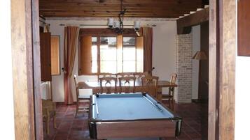 Una habitación con mesa de billar, sillas de madera, pared de ladrillo y ventana con cortinas.