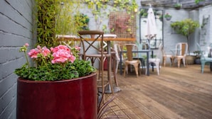 A patio area with a wooden table and chairs, a red planter with pink flowers, and a grey brick wall. - The Loopy Shrew (Shrewsbury)