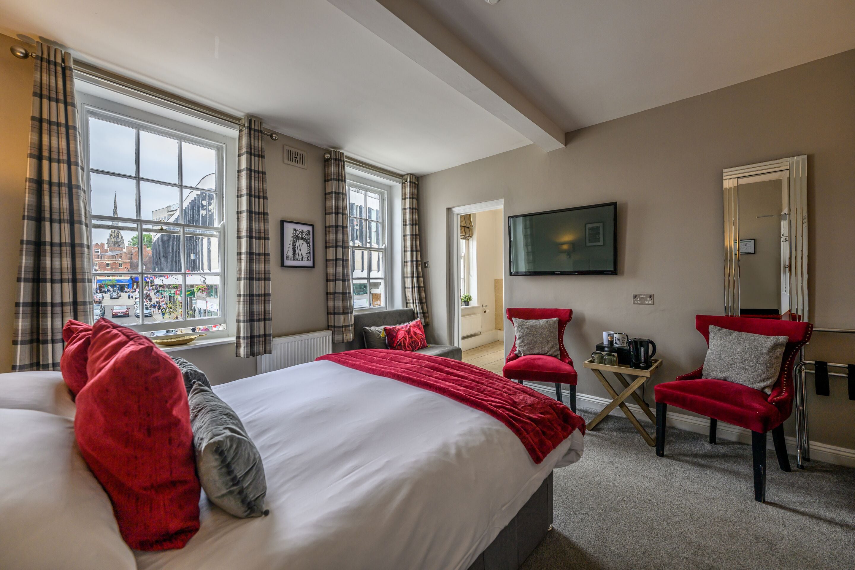 A hotel room with a large bed, two red chairs, a small table with a kettle, a TV, and a view of the city.