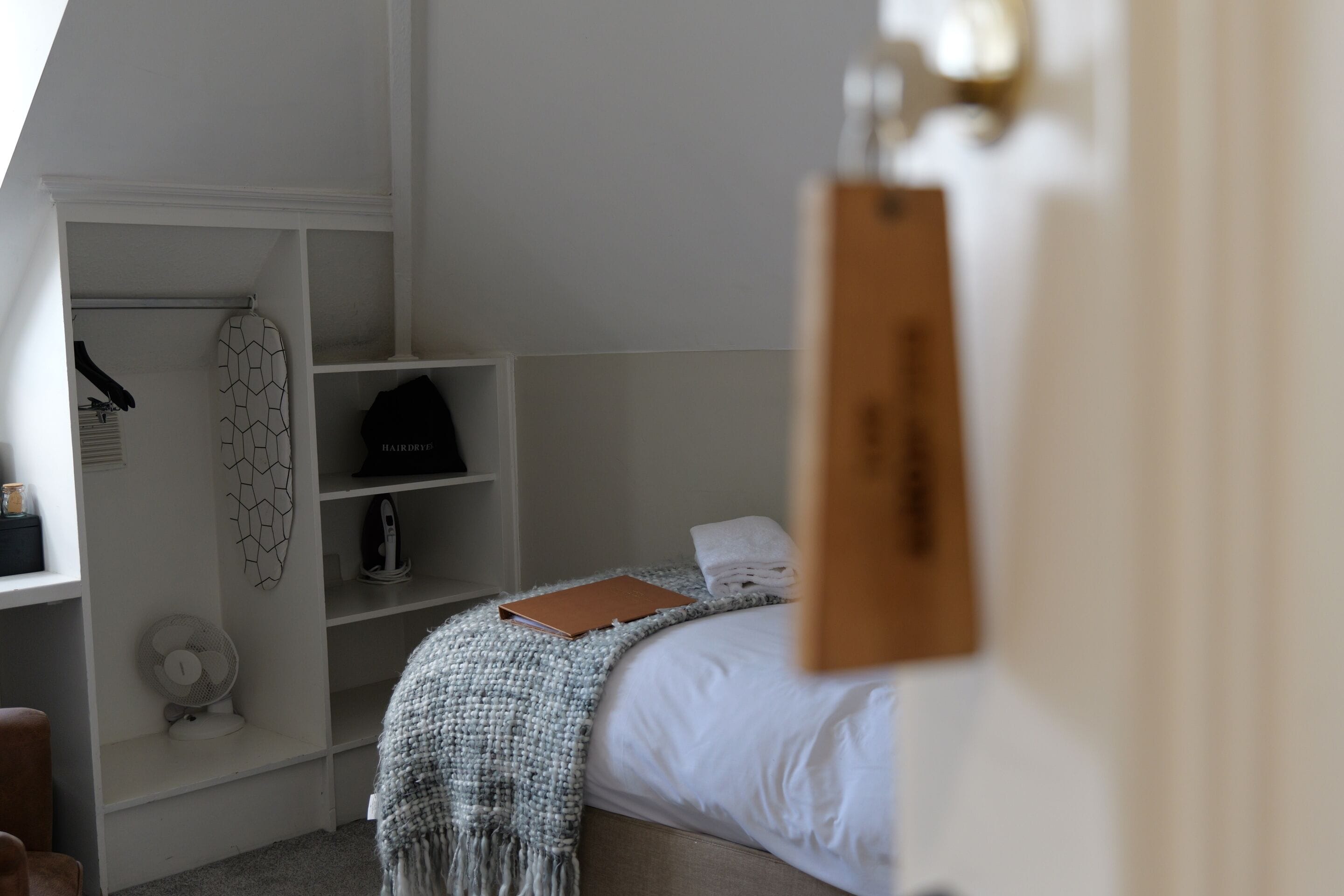 Un dormitorio con una cama, una estantería con un libro, un ventilador y un objeto fijado a la pared.