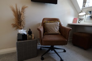 A room with a brown leather chair, a small table with a vase of dried plants, a grey ottoman, and a desk with a lamp. - The Loopy Shrew (Shrewsbury)