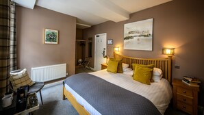 Une chambre à coucher comprenant un lit, des tables de chevet, un radiateur, une chaise, un porte-manteau et une porte donnant sur une autre pièce.